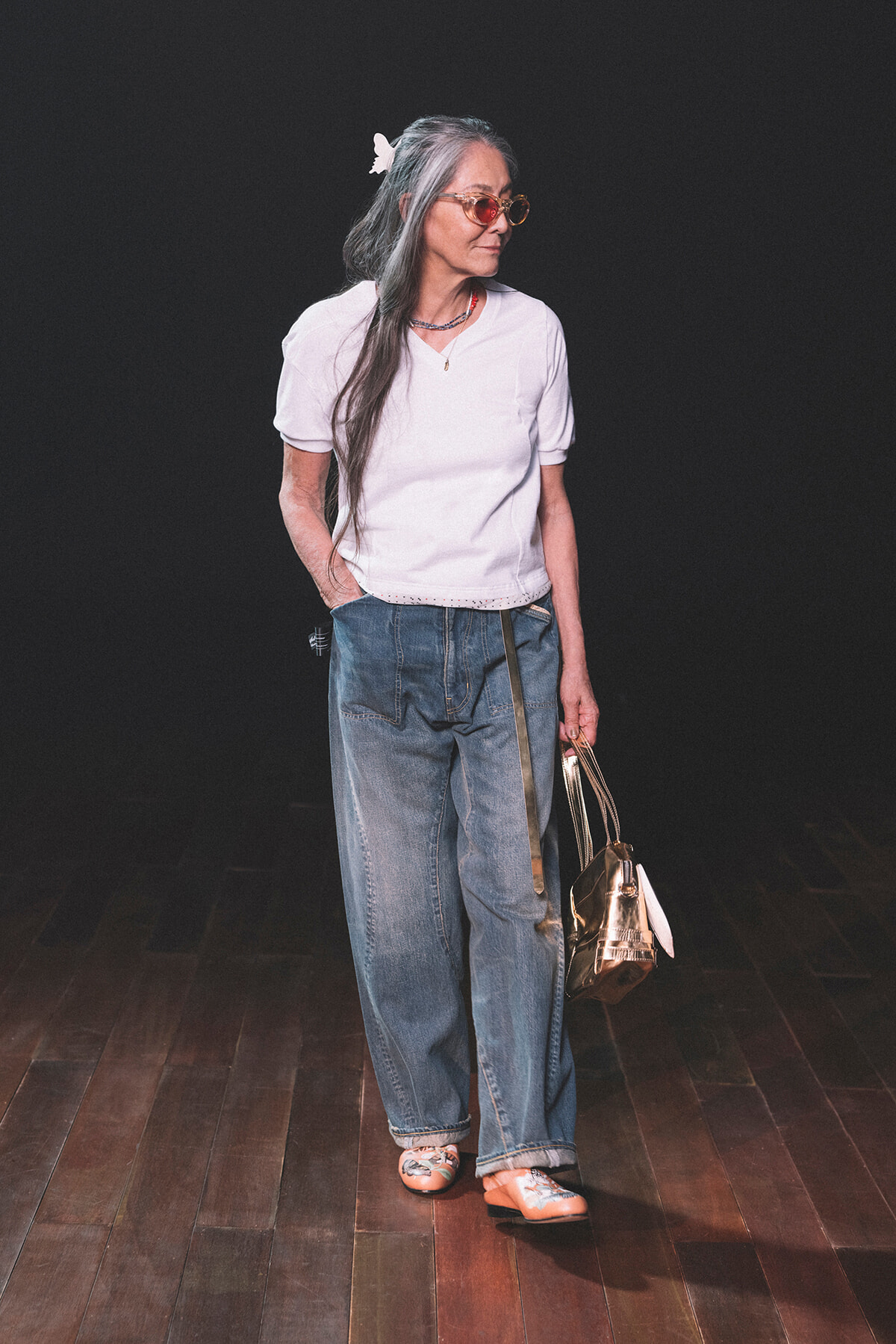 Older woman with gray hair, white top, loose jeans, butterfly hair clip, pink sunglasses, carrying a metallic handbag on a dark stage.