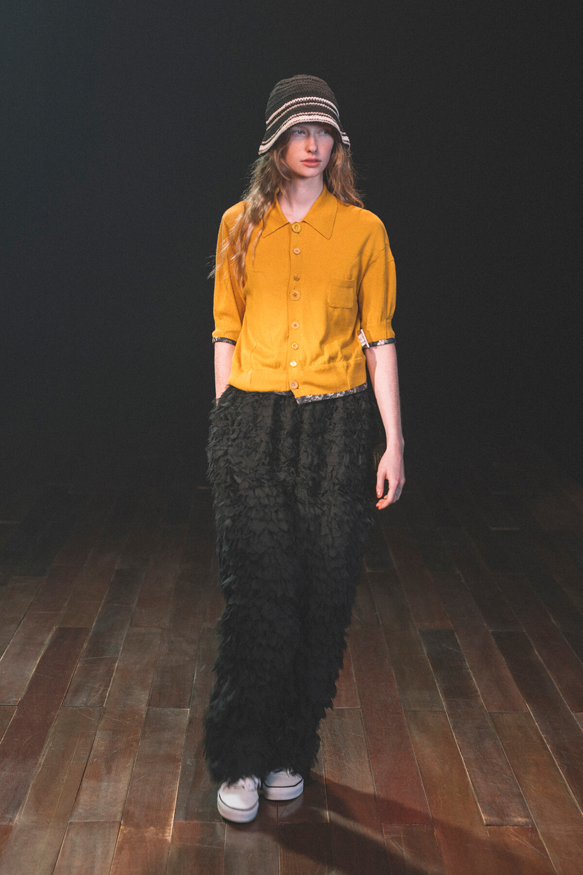 Model on a runway wearing a mustard yellow button-down shirt, black textured wide-leg pants, a striped knit bucket hat, and white sneakers.