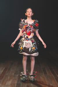 Model on a runway wearing an avant-garde dress covered in colorful knitted textures and floral embellishments.