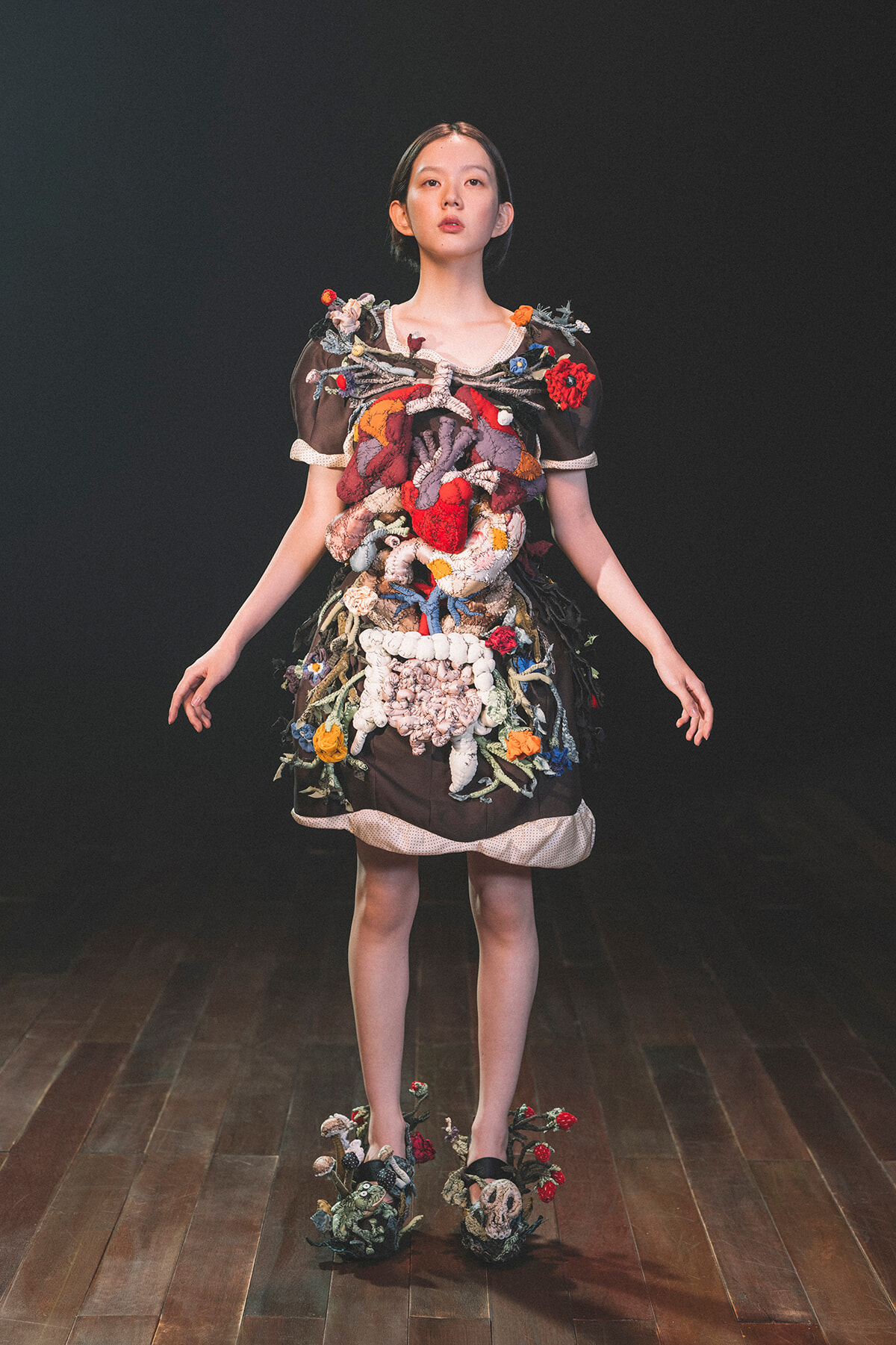 Model on a runway wearing an avant-garde dress covered in colorful knitted textures and floral embellishments.
