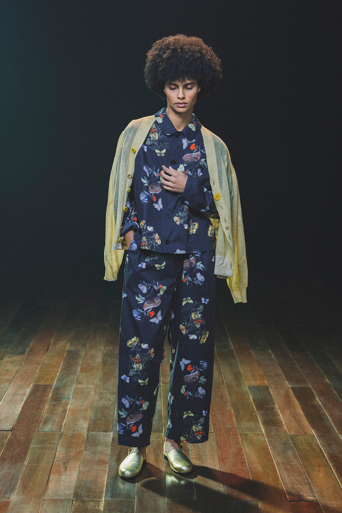 Model in a navy floral suit with a pale yellow cardigan draped over shoulders, standing on a wooden stage against a dark backdrop.