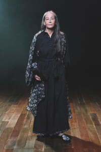 Older woman in a black belted dress with a floral cape, standing on a wooden floor and holding a black clutch against a dark backdrop.