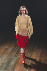 Model walking a runway in a beige cardigan over a sheer beige top, a red feathered skirt, red boots, and pink sunglasses.