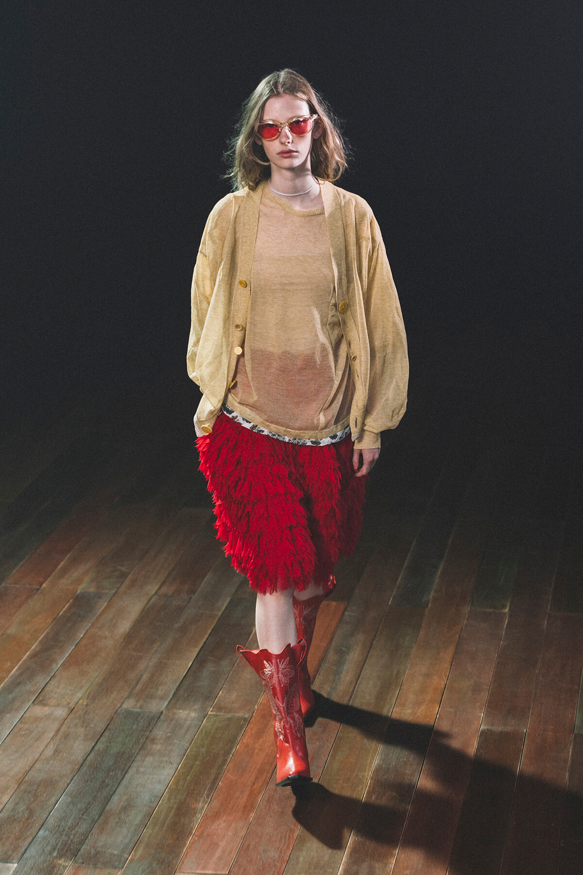 Model walking a runway in a beige cardigan over a sheer beige top, a red feathered skirt, red boots, and pink sunglasses.
