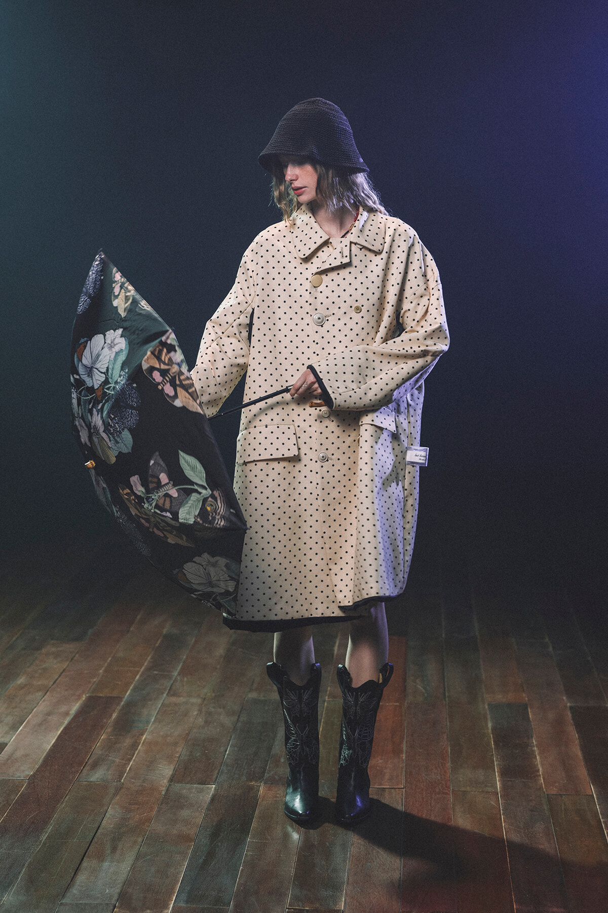 Person wearing a beige polka dot trench coat and bucket hat, holding a floral umbrella indoors on a wooden floor.
