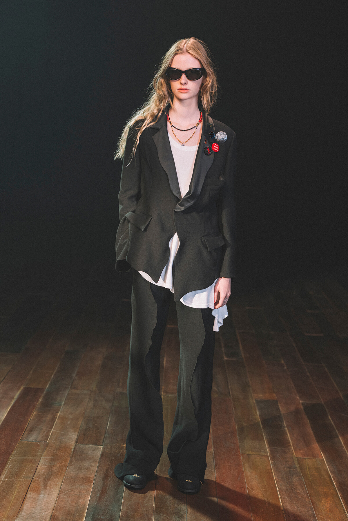 Model on a wooden runway wearing a black oversized blazer, matching wide-leg pants, a white top, sunglasses, and badge pins on the blazer.