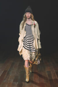 Model walking runway in a striped black-and-white dress under a beige jacket with patches, cream feathered skirt, tan cowboy boots, and a black knit hat, carrying a metallic bag.