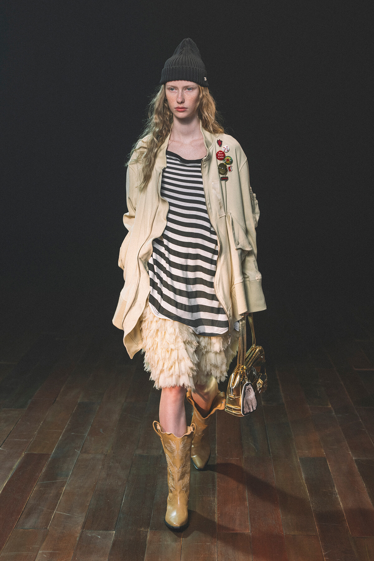 Model walking runway in a striped black-and-white dress under a beige jacket with patches, cream feathered skirt, tan cowboy boots, and a black knit hat, carrying a metallic bag.