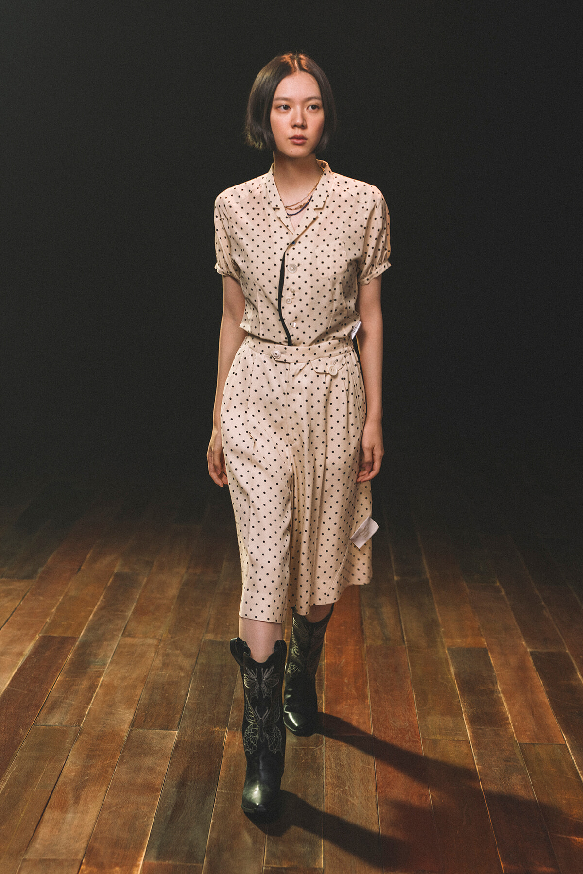 Model walking on a wooden runway wearing a beige polka-dot short-sleeve blouse and matching wide-leg pants with black boots.