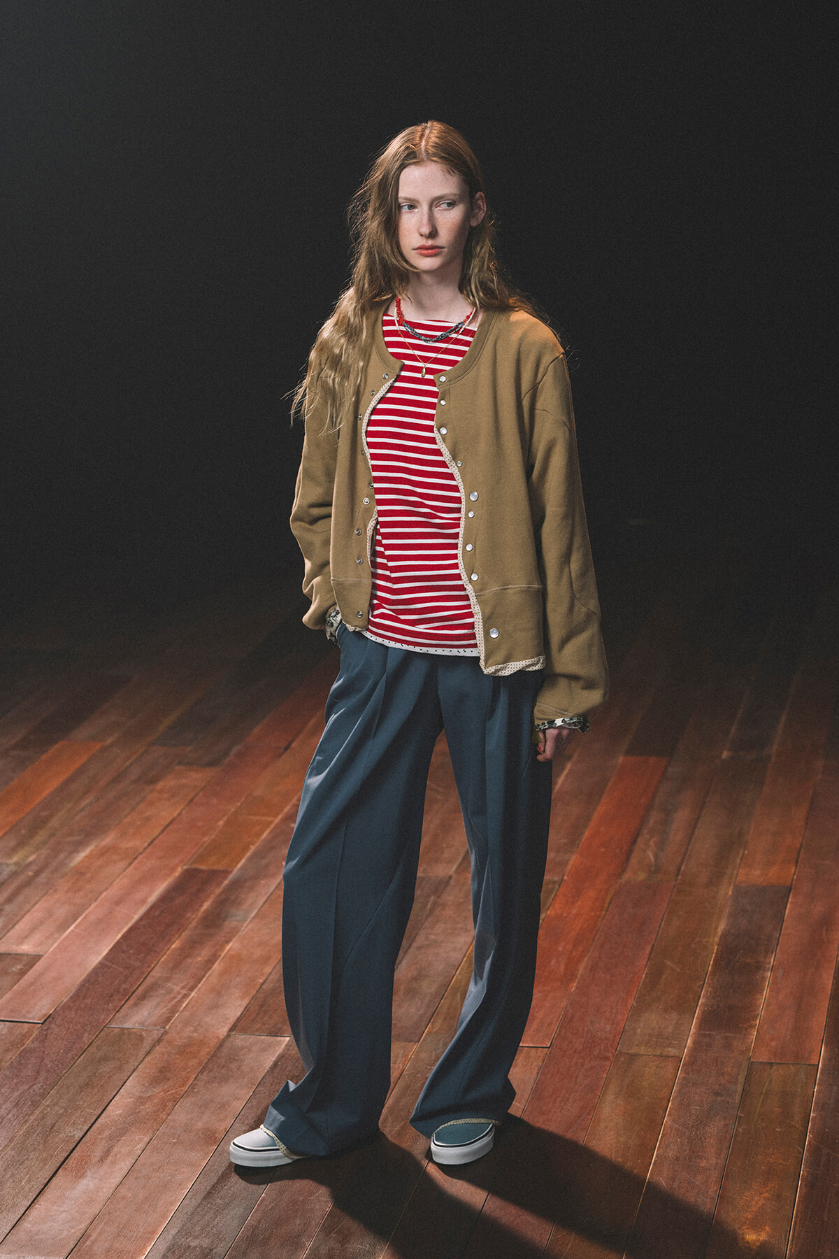 Young woman standing on a wooden floor in a tan jacket, red striped shirt, and blue wide-leg pants against a dark background.