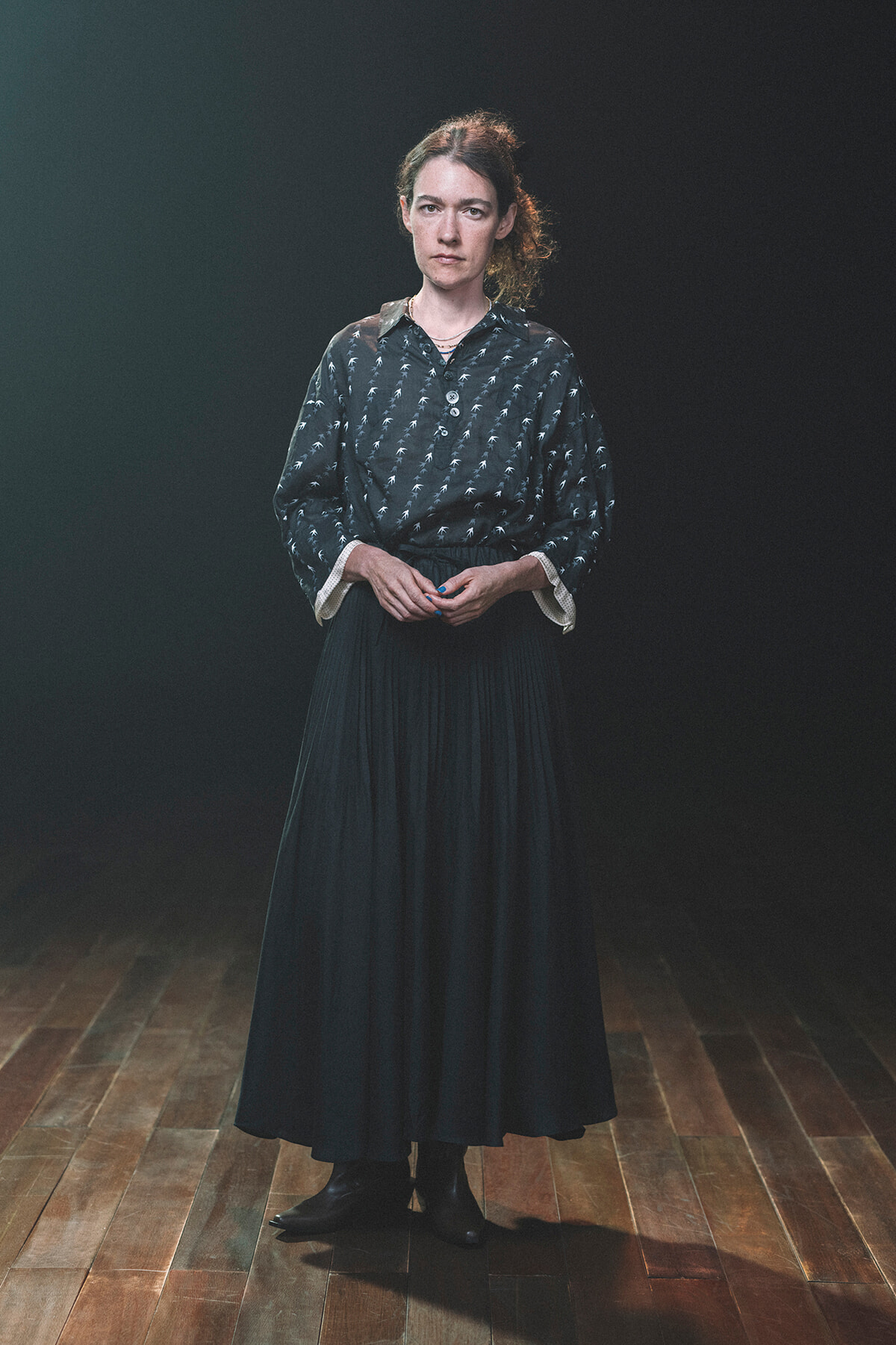 Portrait of a woman standing in a dim studio, wearing a dark patterned blouse and a long black skirt against a dark backdrop.