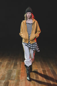 Female model walking on a dark stage, wearing a mustard zip-up hoodie over a black-and-white striped top, white pants, black patterned knee-high boots, and a black knit beanie, on a wooden floor.