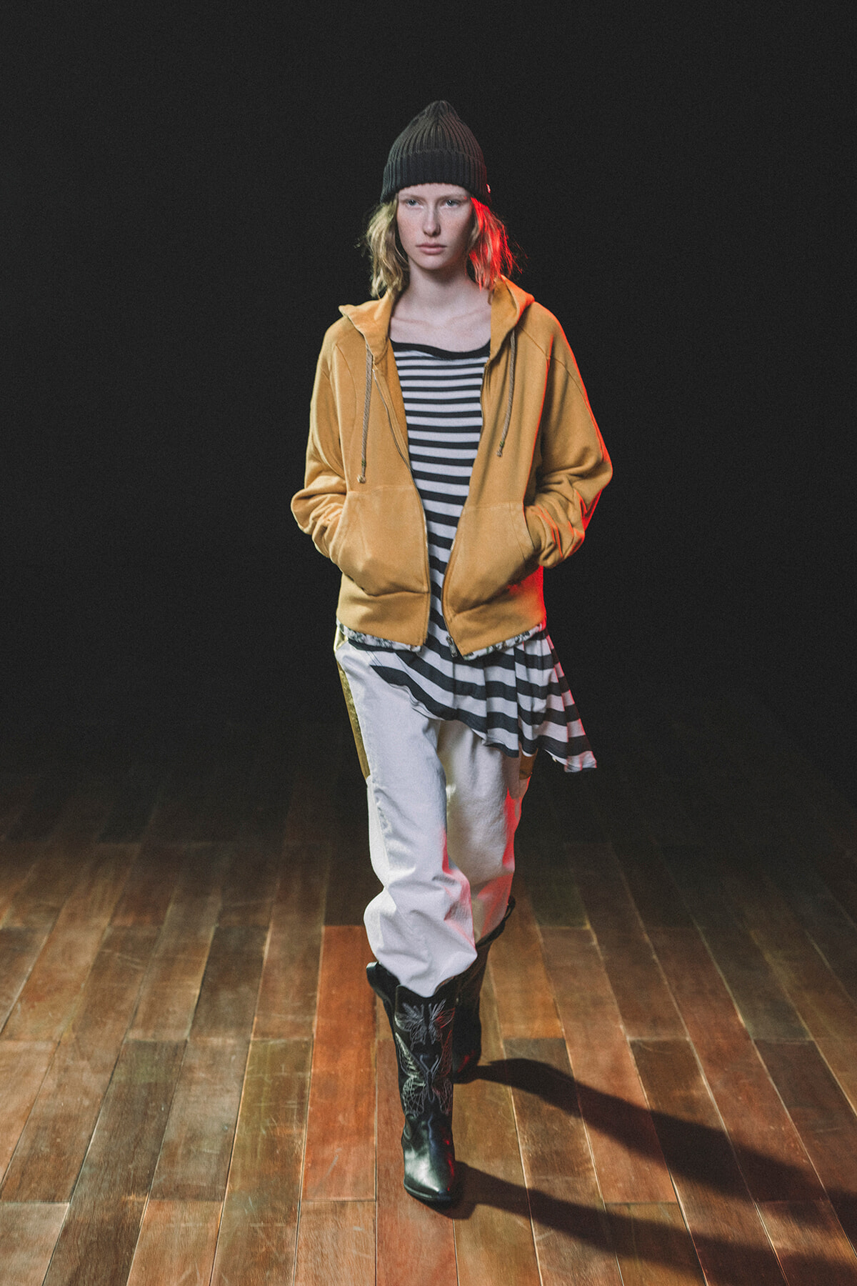 Female model walking on a dark stage, wearing a mustard zip-up hoodie over a black-and-white striped top, white pants, black patterned knee-high boots, and a black knit beanie, on a wooden floor.