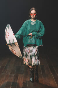 Model in a green layered outfit with a printed skirt, red-tinted sunglasses, and knee-high boots, holding a graphic umbrella in a dark studio.