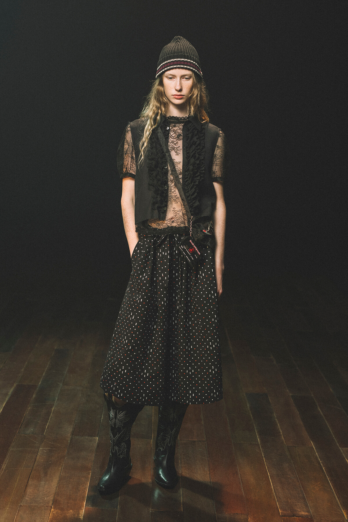 Model wearing a sheer lace top with a black vest over it, a black polka-dot midi skirt, tall boots, and a knit beanie, standing on a wooden floor against a dark background.