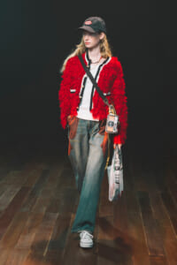 Model walking a runway in a red textured shaggy jacket over a white top, with a crossbody bag and bag hanging from the hip, wearing a cap and white sneakers.