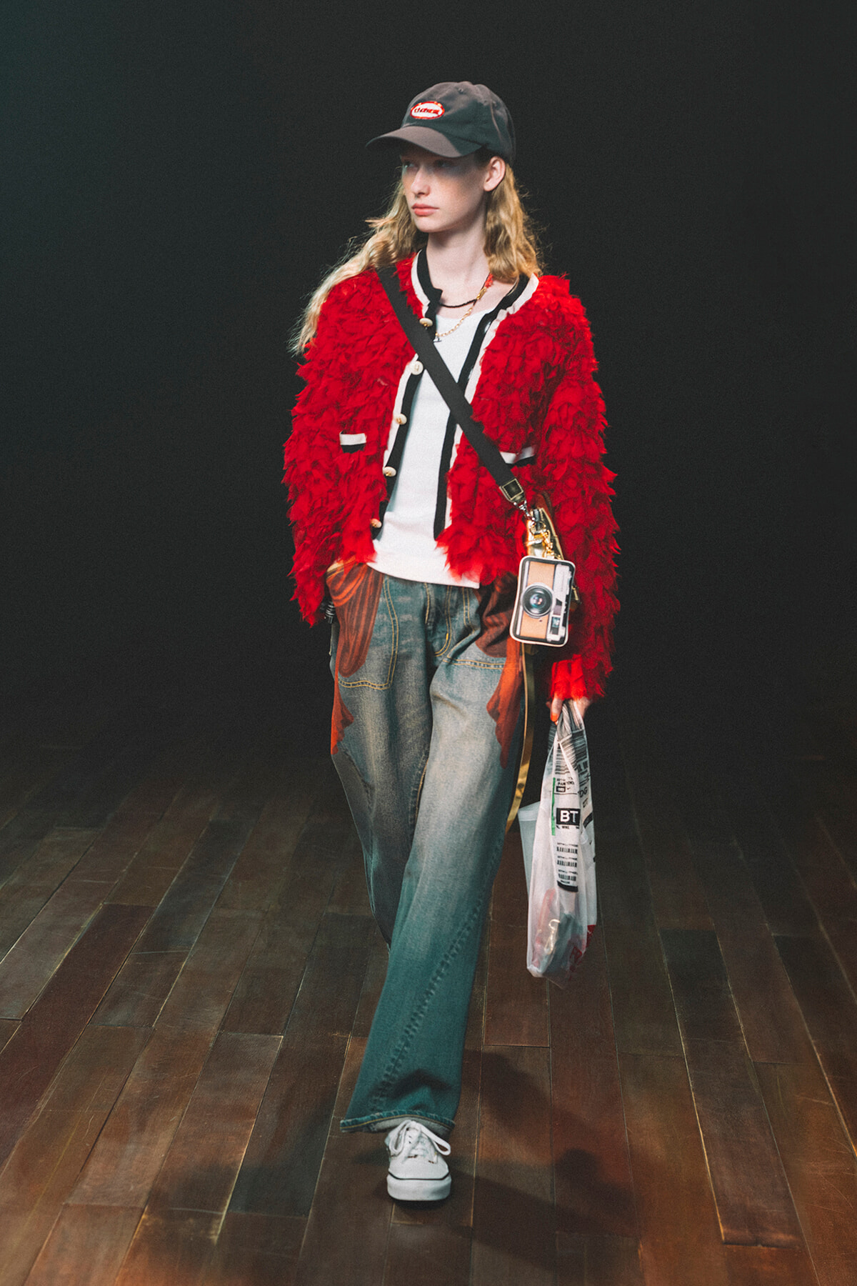 Model walking a runway in a red textured shaggy jacket over a white top, with a crossbody bag and bag hanging from the hip, wearing a cap and white sneakers.