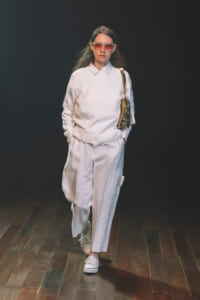 Model walking the runway in an all-white outfit: knit sweater, wide-leg pants, white sneakers, pink-tinted sunglasses, and a beige shoulder bag.