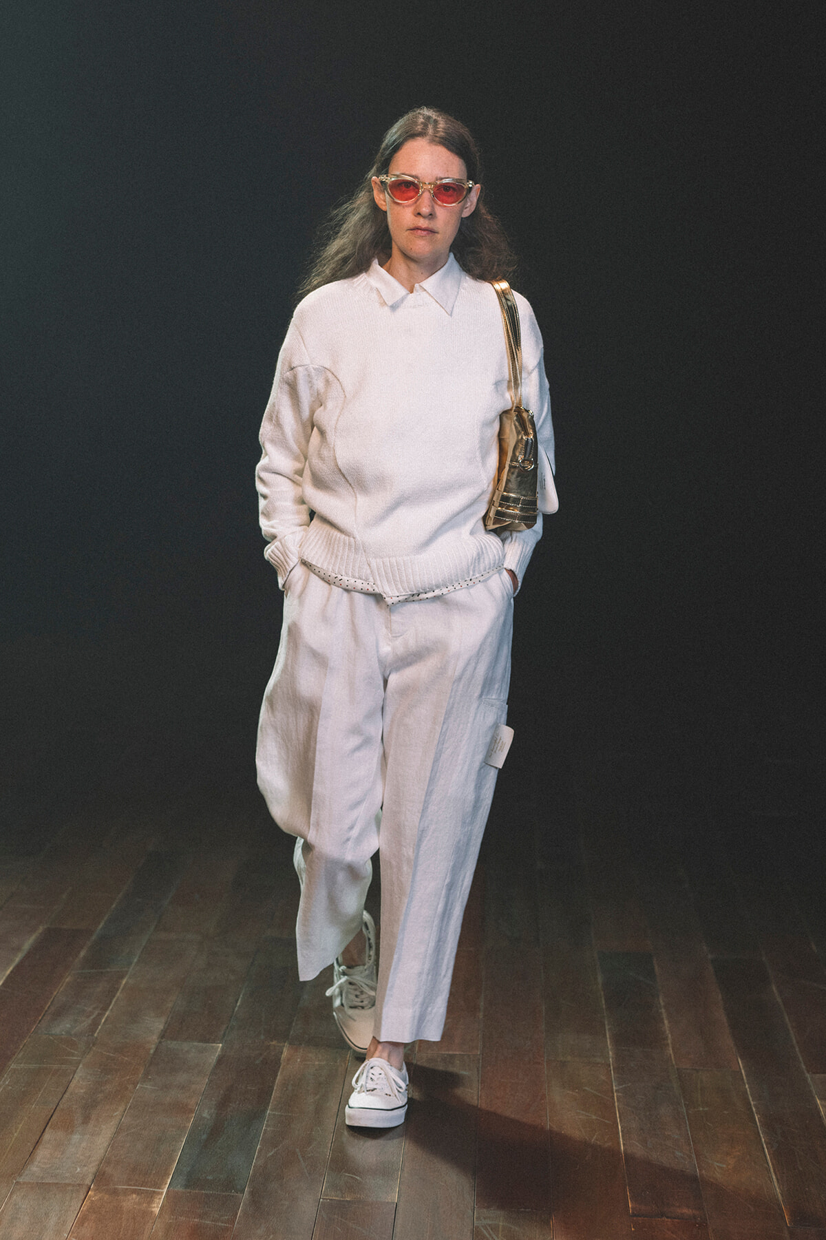 Model walking the runway in an all-white outfit: knit sweater, wide-leg pants, white sneakers, pink-tinted sunglasses, and a beige shoulder bag.