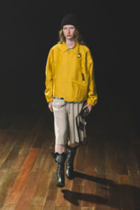 Model wearing a bright yellow jackets over beige shorts, black beanie, and black embroidered boots on a wooden runway floor.