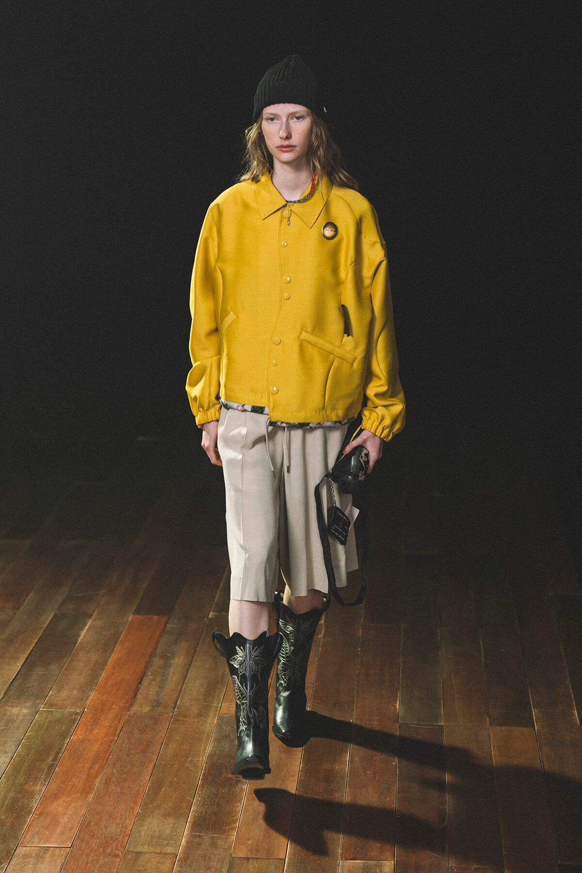 Model wearing a bright yellow jackets over beige shorts, black beanie, and black embroidered boots on a wooden runway floor.
