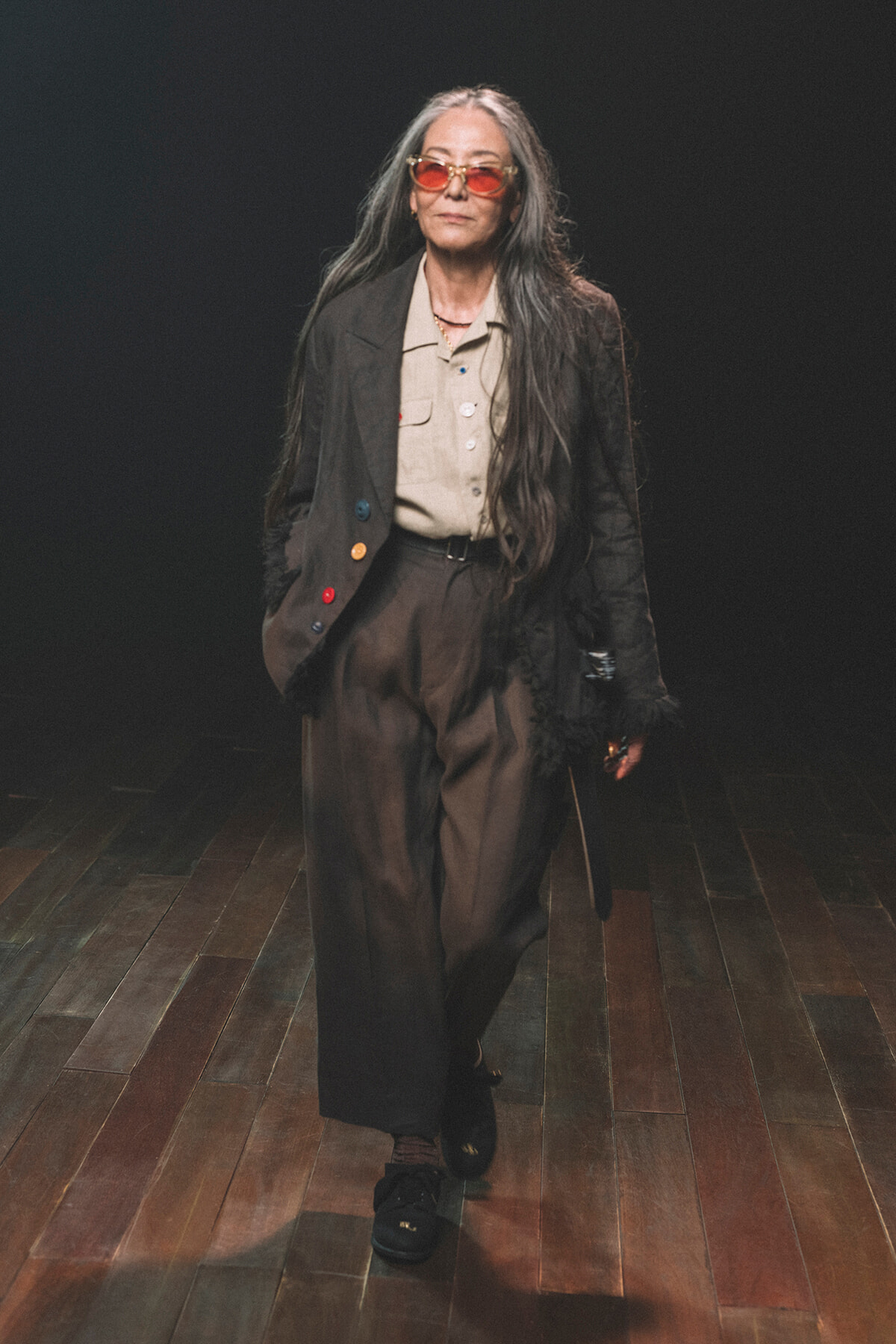 Older woman with long gray hair wearing a brown suit and red-tinted sunglasses, walking on a dark stage with wooden floor.