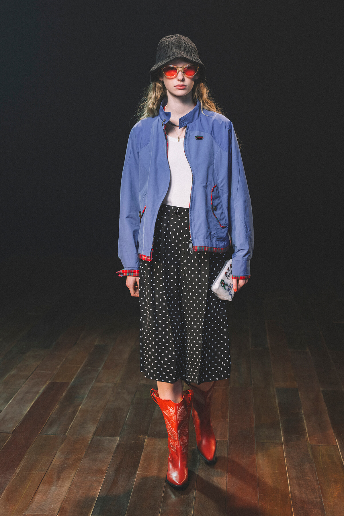Model wearing a blue zip jacket over a white top, black polka-dot skirt, red boots, red sunglasses, and a bucket hat on a dark backdrop.