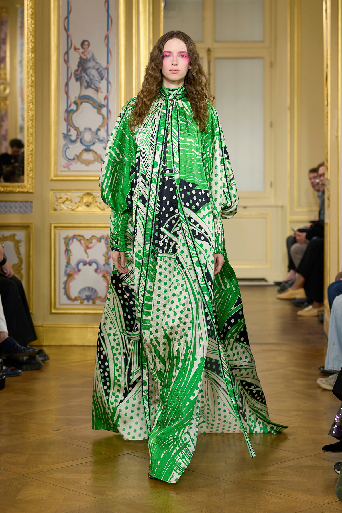 Model on a fashion runway wearing a green abstract-print, floor-length gown with voluminous sleeves, in a gilded ornate room background.