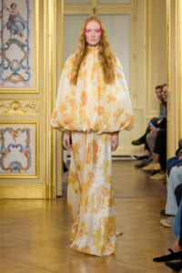 Model on a runway wearing a sparkling peach-floral cape gown with a voluminous cape and long skirt, in a gilded, ornate room.