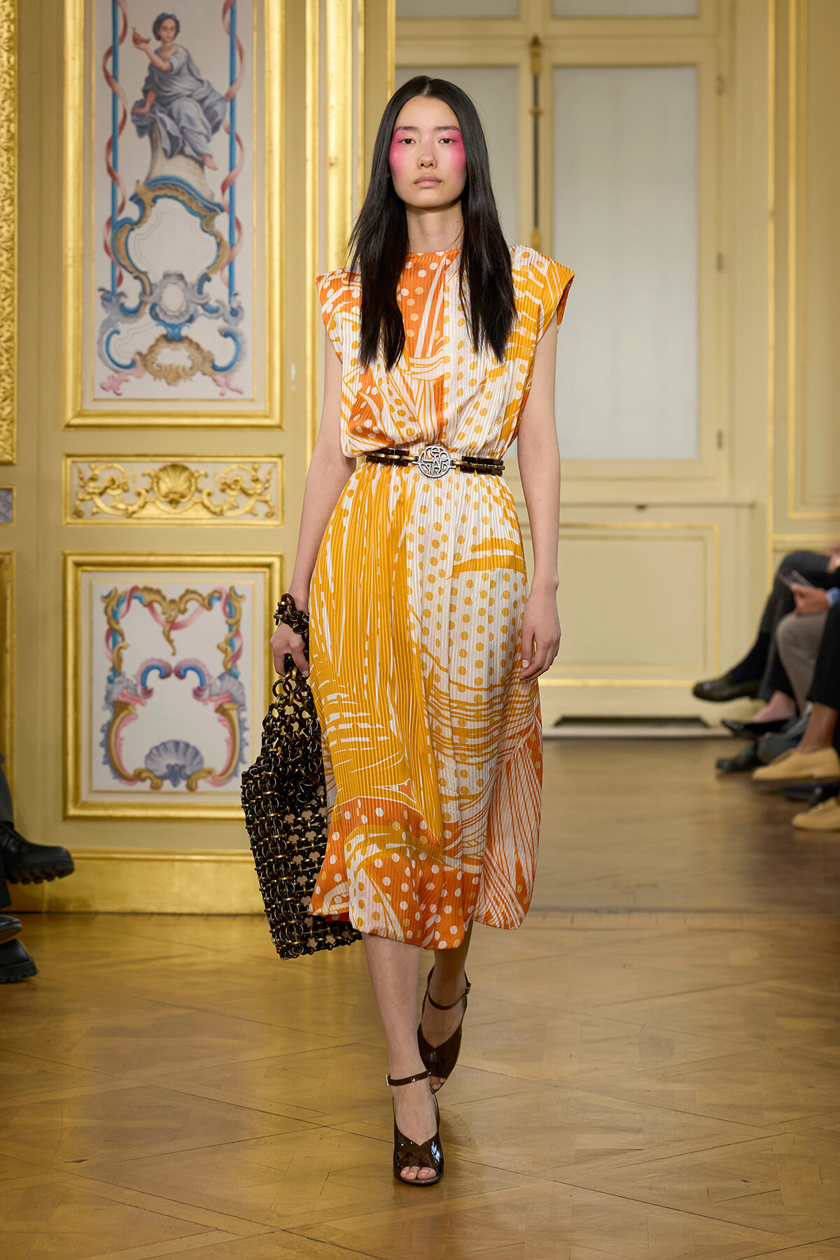 Model walking a runway in a bright orange and white patterned midi dress with a black belt, holding a black woven handbag, in a gilded, ornate room.