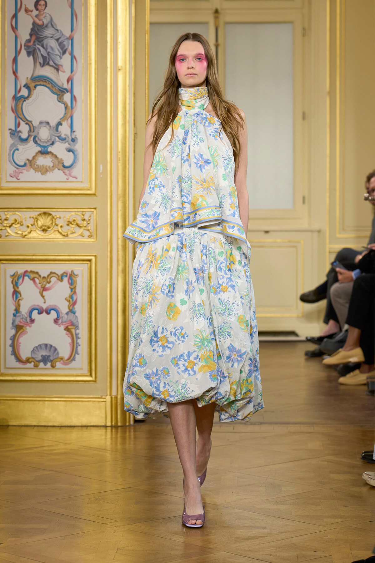 Model walks the runway in a pastel blue floral gown with a layered, voluminous skirt in a gilded, ornate room.