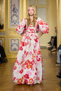 Model walking runway in a white dress with bold pink and red floral print and oversized puff sleeves in a gilded room setting.