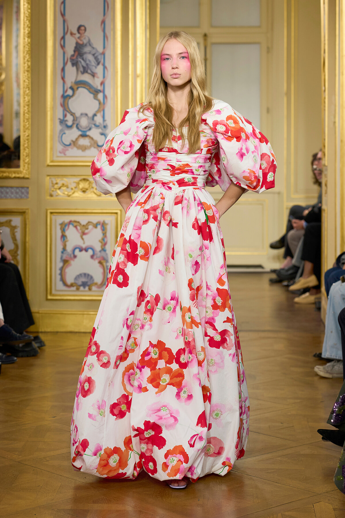 Model walking runway in a white dress with bold pink and red floral print and oversized puff sleeves in a gilded room setting.