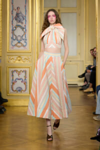 Model on a runway wearing a pastel striped two-piece outfit with a large bow at the shoulder in a gilded, ornate room