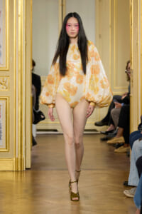 Runway model in a glittery gold floral one-piece with dramatic balloon sleeves on a gilded catwalk backdrop.