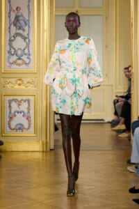 Model in pastel floral dress with a tied waist and puff sleeves walking a gilded runway.