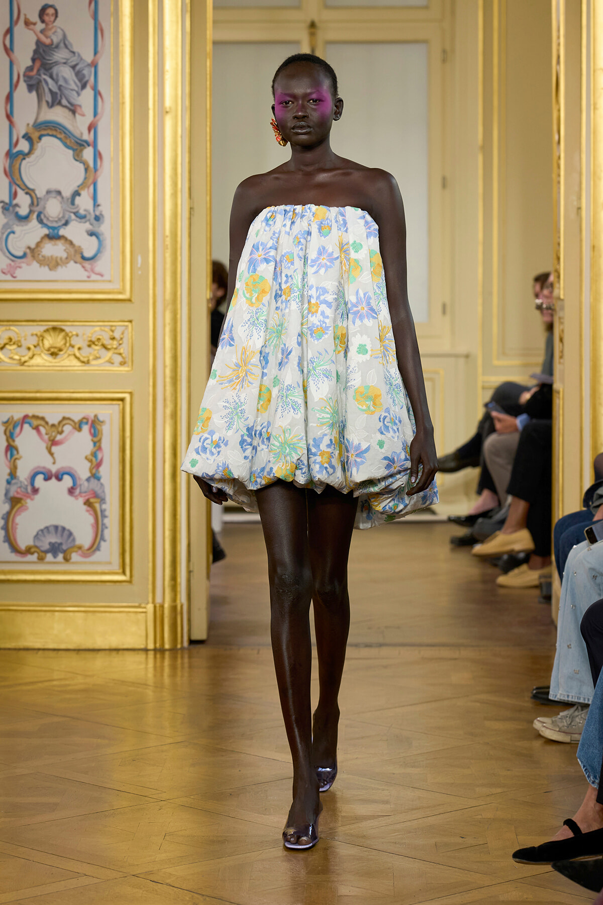 Model walks a pastel floral strapless bubble dress down a runway in a gilded, ornate room.