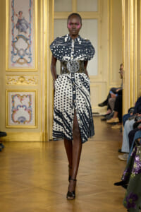 Runway model in a sculptural black-and-white print dress with a wide belt, walking in a gilded, ornate room through a fashion show.