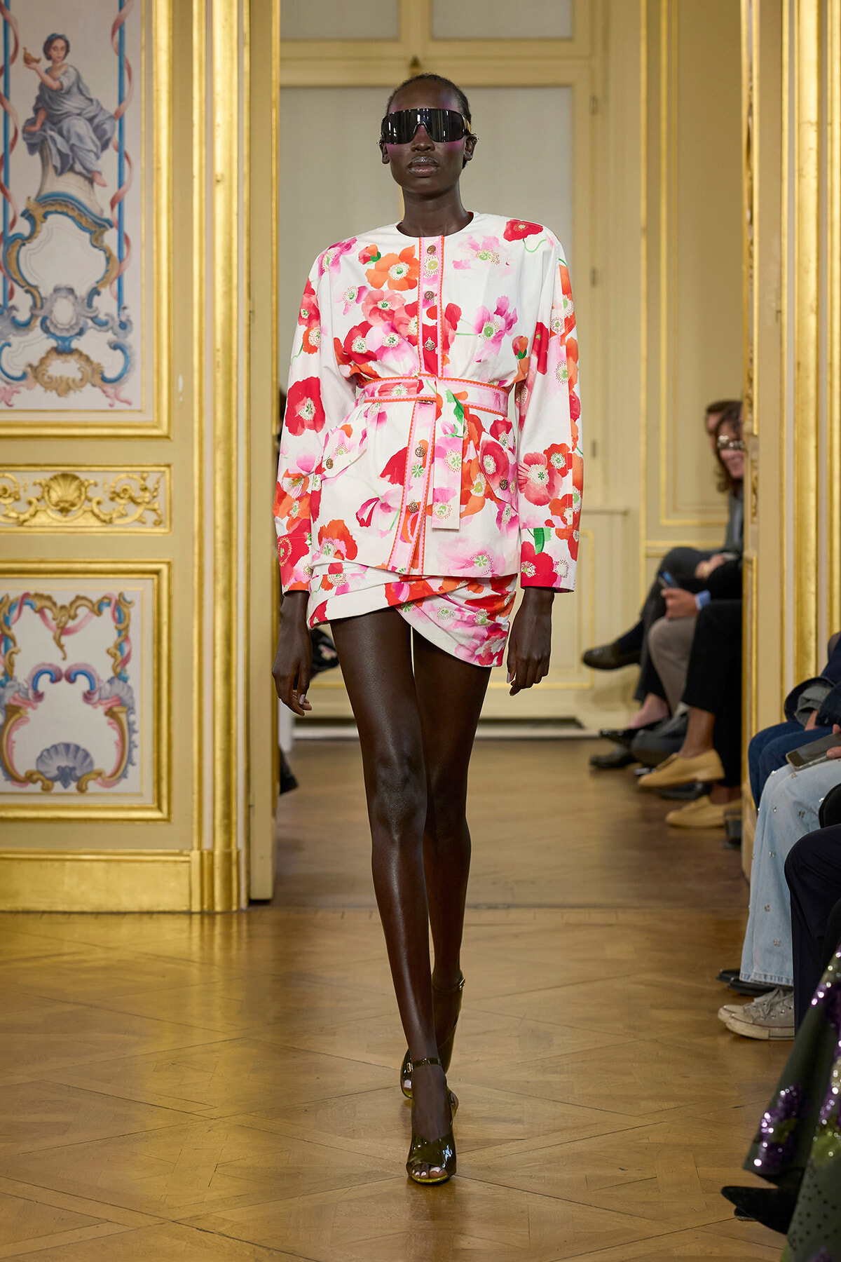 Model walking a fashion runway in a floral pink-and-white dress with a belt, dark sunglasses, and high-heeled sandals in a gilded ornate room.