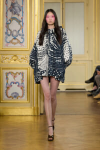 Female model walking a runway in a loose black-and-white geometric-patterned tunic with billowy sleeves in a gilded, ornate room outlinework backdrop.