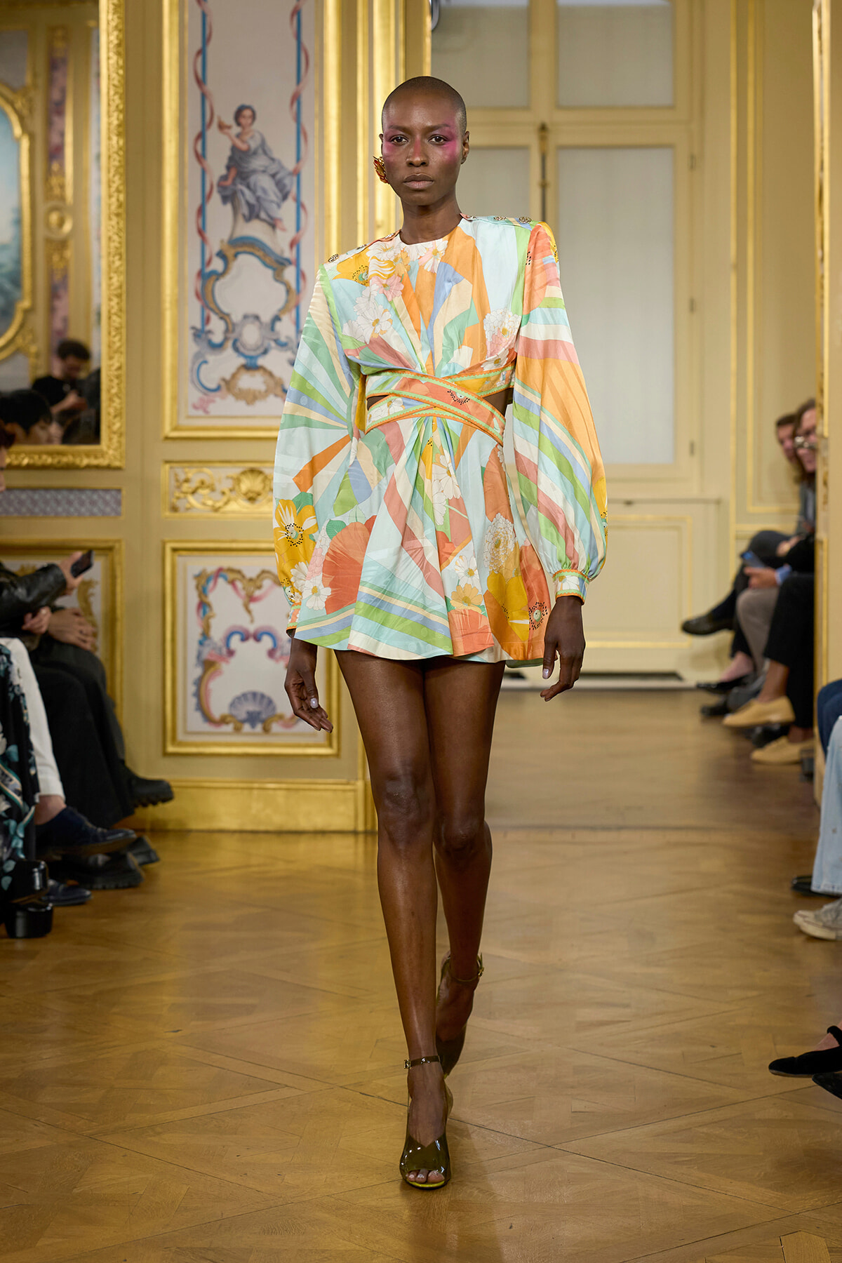 Model walks a runway in a pastel striped floral mini dress with a wrapped belt, wearing olive heels in a gilded rococo ballroom.