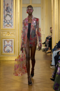 Model walks the runway wearing a translucent pink floral blazer over a black bodysuit, carrying a matching clear tote bag.