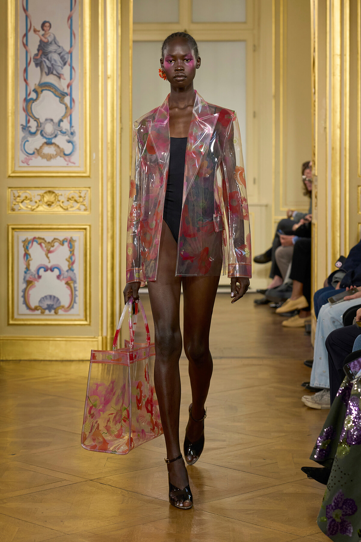 Model walks the runway wearing a translucent pink floral blazer over a black bodysuit, carrying a matching clear tote bag.