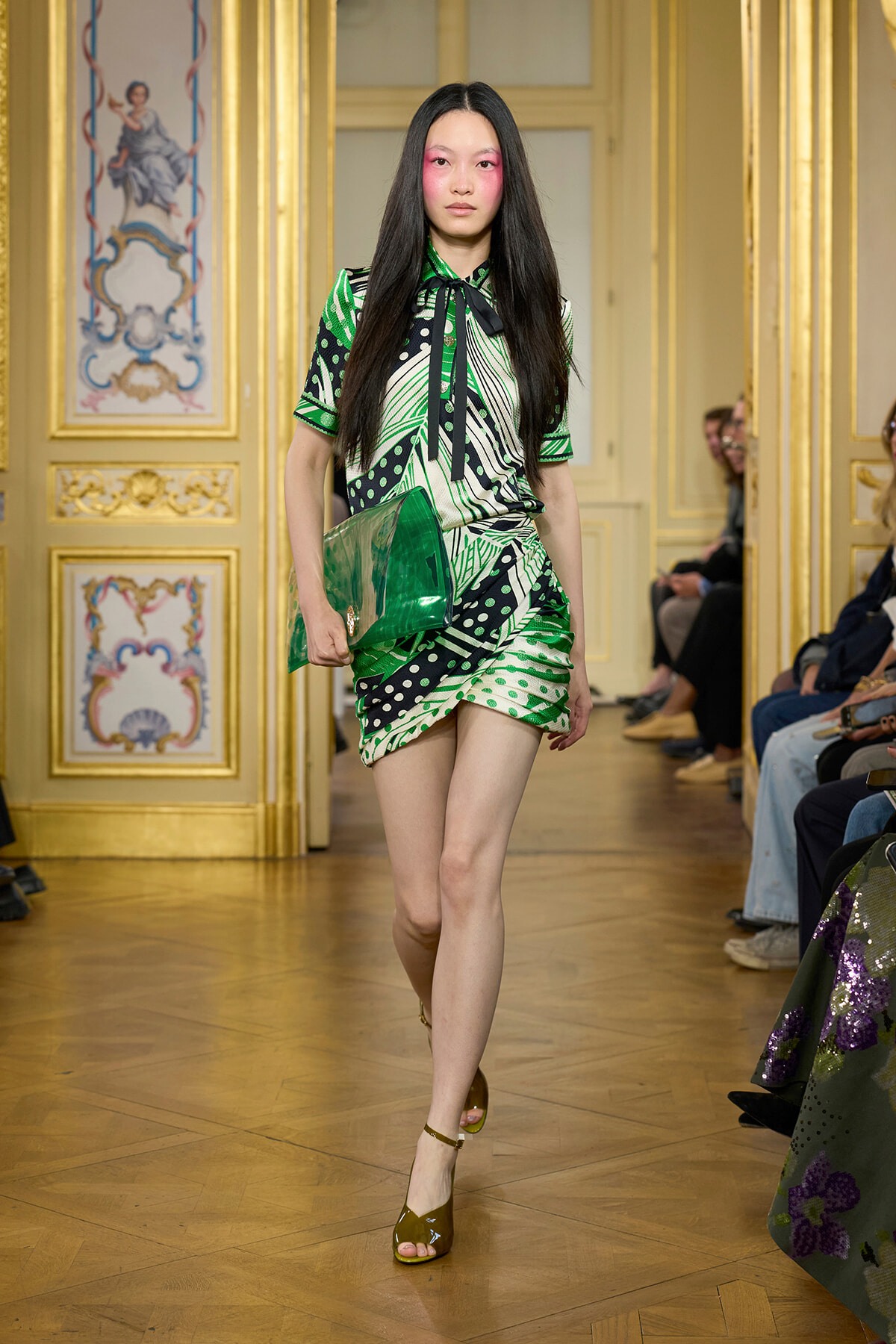 Model walks the runway in a green geometric dress with a black bow, carrying a shiny green clutch in an ornate gold room.