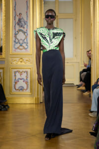 Model walks a runway in a navy floor-length skirt and a green patterned top, wearing large sunglasses in a gilded, ornate room.