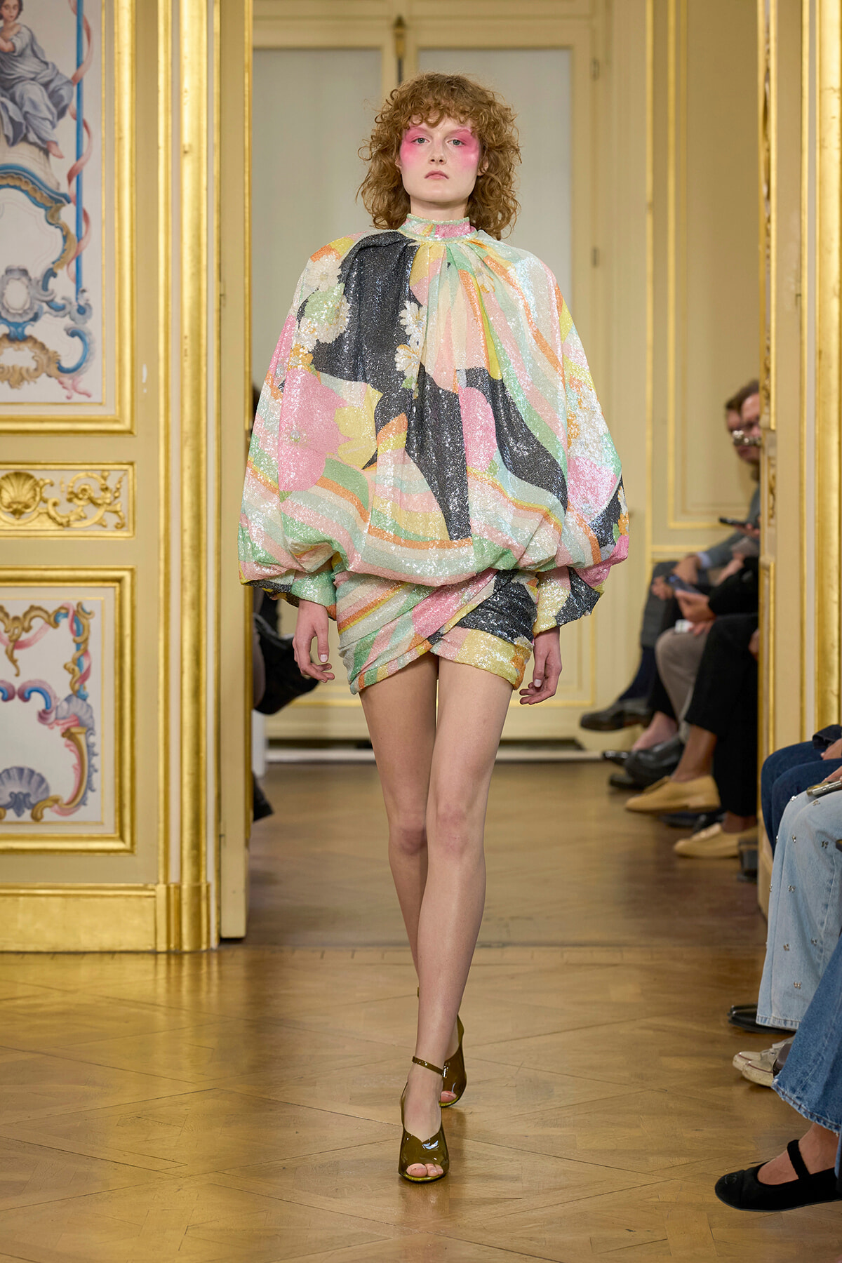 Model walking a fashion runway in a voluminous, iridescent pastel dress with pink makeup in an ornate gold-trimmed venue.