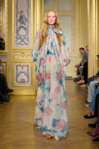 Model walks a runway in a floor-length sheer floral gown with pastel pink and blue blooms in a gilded, ornate room.