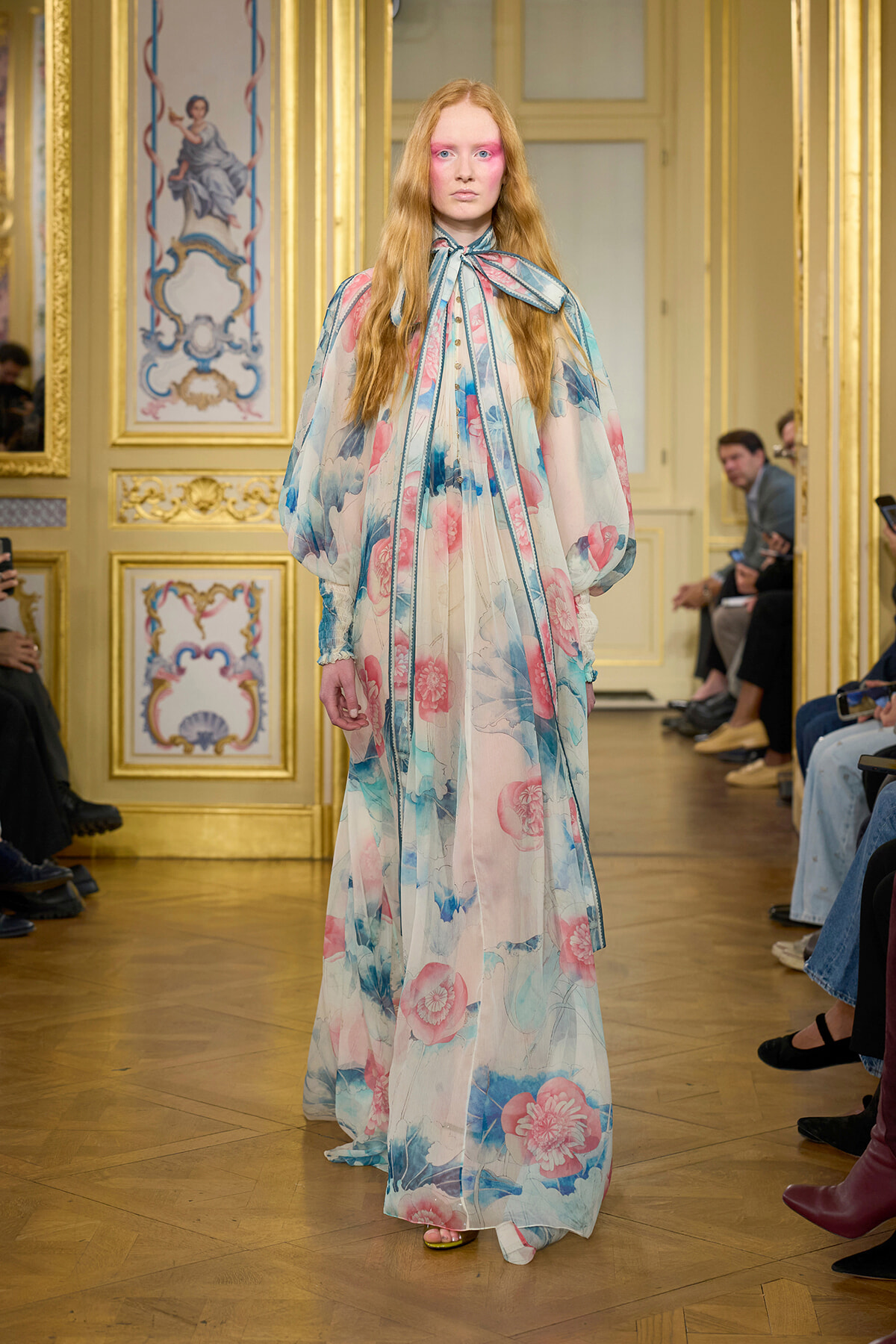 Model walks a runway in a floor-length sheer floral gown with pastel pink and blue blooms in a gilded, ornate room.