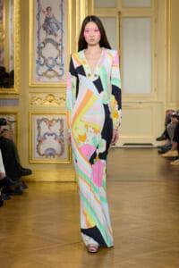 Model walking the runway in a long pastel-colored geometric print dress against a gilded ornate wall backdrop.