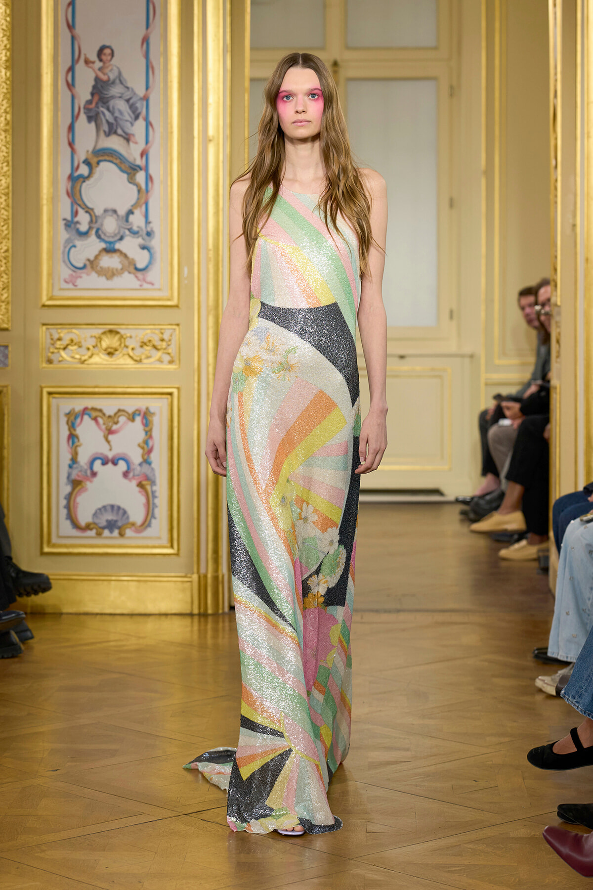 Model walking the runway in a long pastel, iridescent geometric gown in a gilded, ornate room with decorative panels.